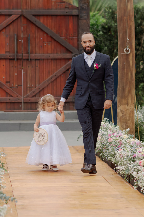 Noivo Matheus caminha sorridente pelo corredor da cerimônia ao ar livre de mãos dadas com uma daminha de honra no Boulevard Jacareí. Fotografia de casamento capturando a doçura e alegria do momento, registrada por Gilson Dias.