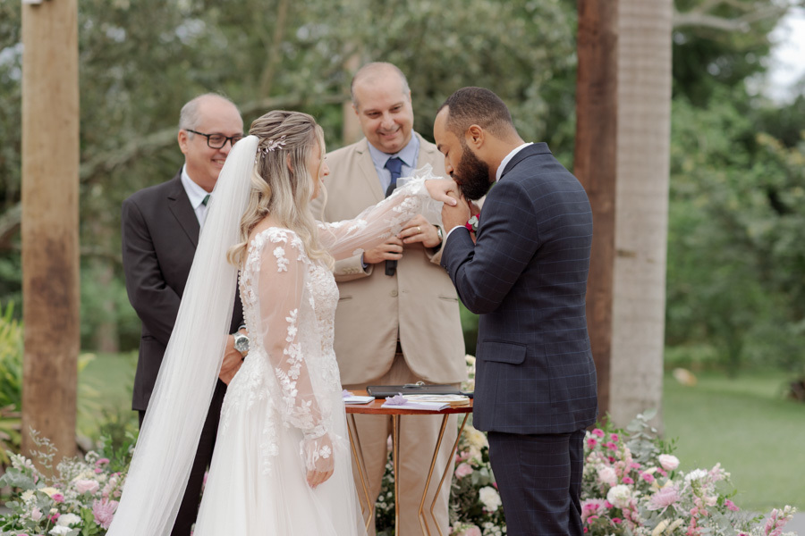 Noivo Matheus beija a mão da noiva Claudia após a troca de alianças no altar do Boulevard Jacareí. O celebrante e o pai da noiva sorriem ao fundo em momento de carinho na cerimônia ao ar livre, registrado pelo fotógrafo Gilson Dias.