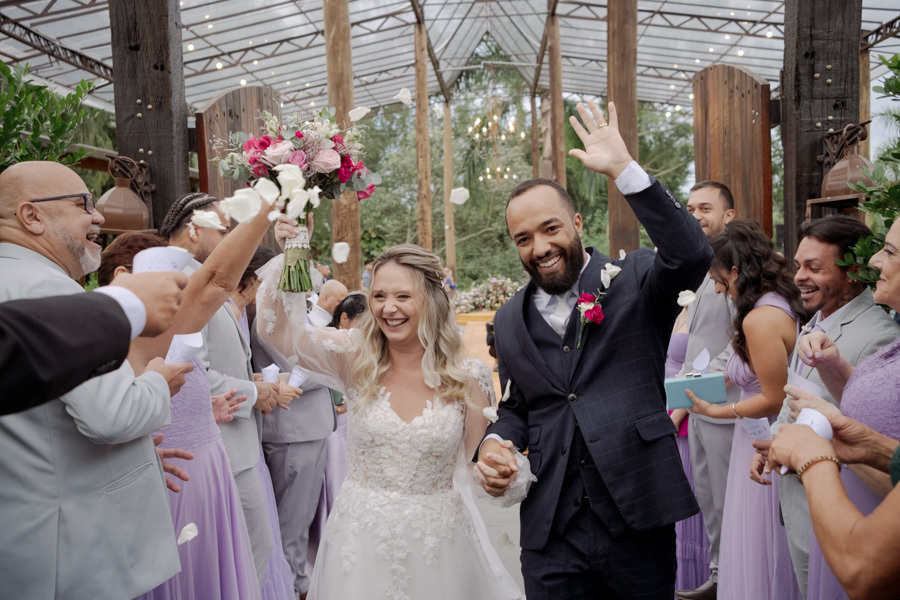 Noivos Claudia e Matheus saem radiantes da cerimônia no Boulevard Jacareí sob chuva de pétalas. Fotografia de casamento celebrando a felicidade do casal de mãos dadas entre os padrinhos, registrada com alegria pelo fotógrafo Gilson Dias.