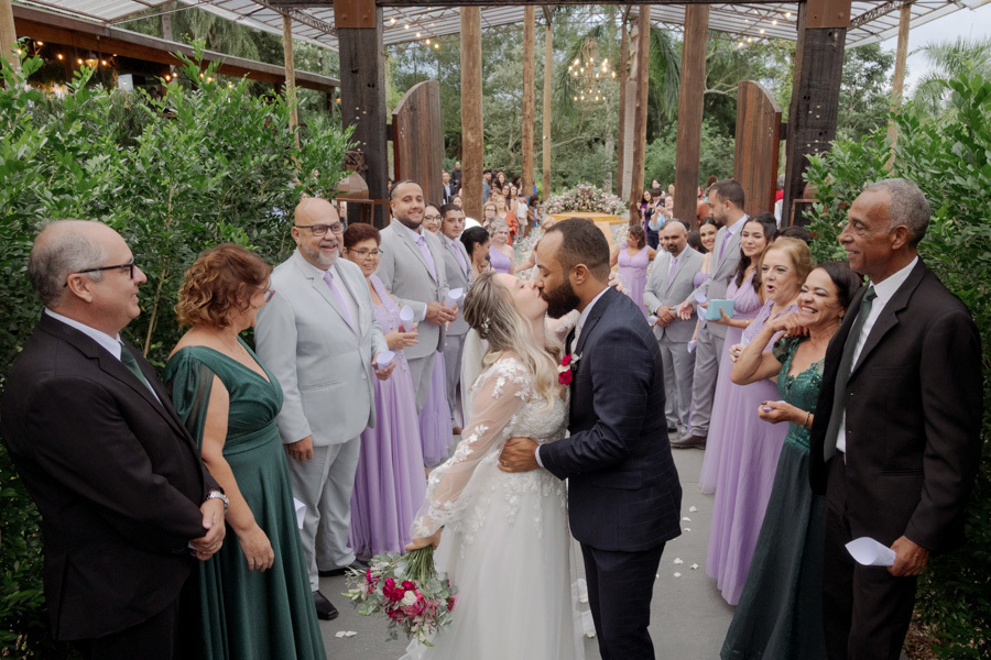 Noivos Claudia e Matheus se beijam apaixonados ao saírem da cerimônia no Boulevard Jacareí. Padrinhos e familiares sorriem e celebram a união do casal no corredor decorado, em fotografia de casamento emocionante registrada por Gilson Dias.