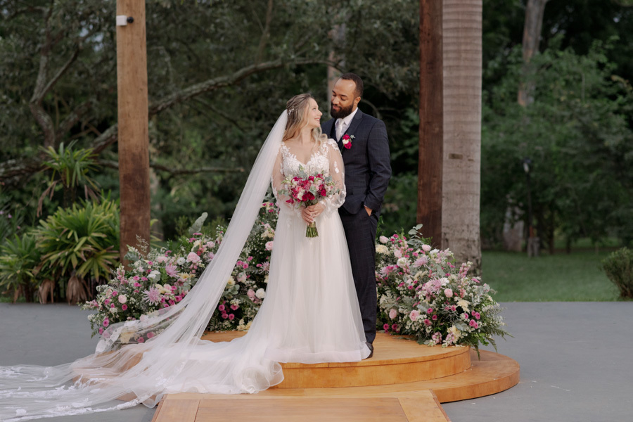 Noivos Claudia e Matheus posam no altar circular do Boulevard Jacareí após a cerimônia. Retrato romântico do casal em plano inteiro, destacando o vestido da noiva com longo véu e o buquê de flores, registrado pelo fotógrafo Gilson Dias.