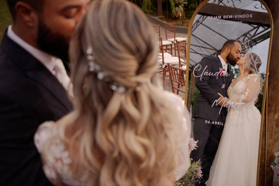 Reflexo dos noivos Claudia e Matheus se beijando em espelho decorativo com seus nomes e data do casamento. Fotografia criativa no Boulevard Jacareí destacando o romantismo e os detalhes da recepção, registrada por Gilson Dias.