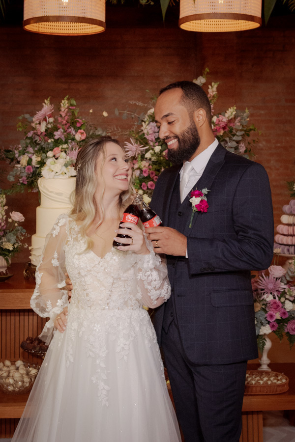 Noivos Claudia e Matheus brindam com garrafas de Coca-Cola em frente ao bolo de casamento no Boulevard Jacareí. Fotografia divertida e espontânea capturando a alegria do casal na mesa de doces, registrada pelo fotógrafo Gilson Dias.