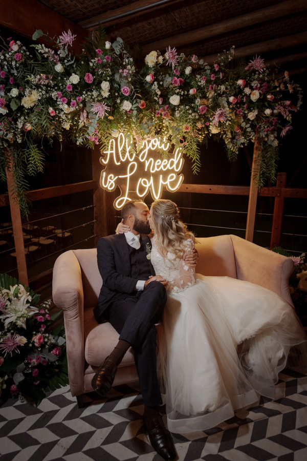 Noivos Claudia e Matheus se beijam em sofá rosa sob letreiro de neon 