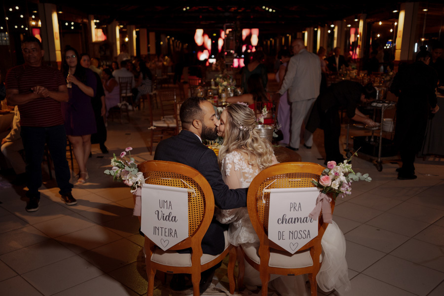 Noivos Claudia e Matheus se beijam sentados à mesa da recepção no Boulevard Jacareí. As cadeiras decoradas com flores trazem as frases 