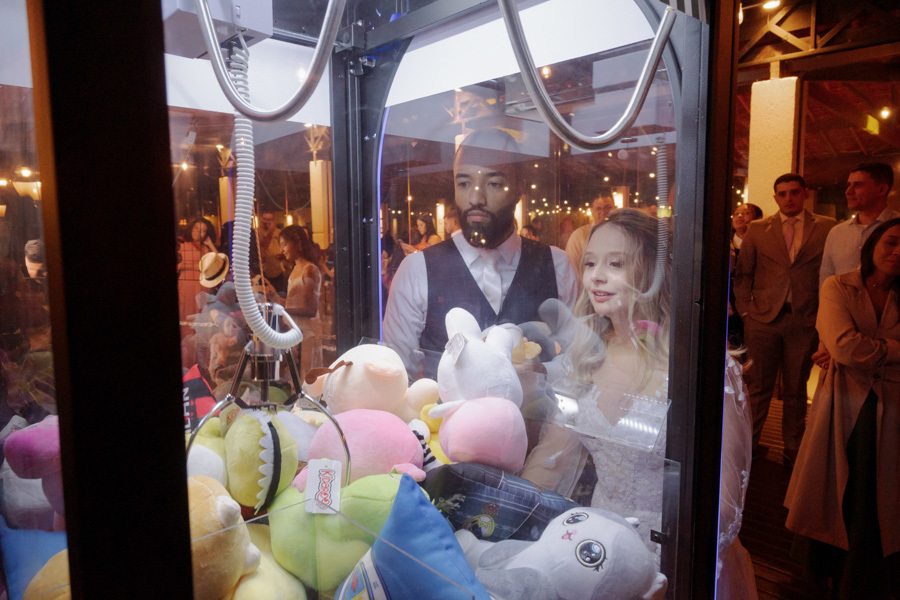 Noivos Claudia e Matheus se divertem tentando pegar um urso de pelúcia em uma máquina de garra durante a festa no Boulevard Jacareí. Momento descontraído e espontâneo da recepção de casamento, registrado pelo fotógrafo Gilson Dias.
