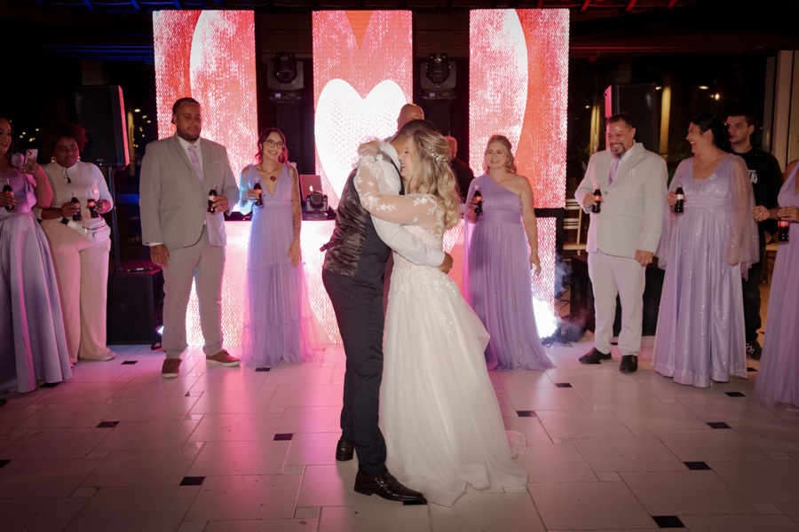 Noivos Claudia e Matheus se abraçam emocionados na pista de dança do Boulevard Jacareí. Padrinhos observam o casal em frente a um painel de LED com corações, celebrando a união em fotografia de festa de casamento registrada por Gilson Dias.