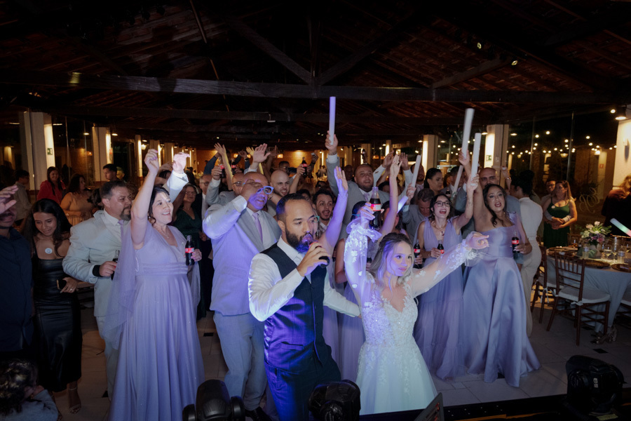 Noivos Claudia e Matheus animam a pista de dança com microfone e bastões de LED no Boulevard Jacareí. Convidados celebram ao fundo em uma festa de casamento vibrante e cheia de energia, registrada pelo fotógrafo Gilson Dias.