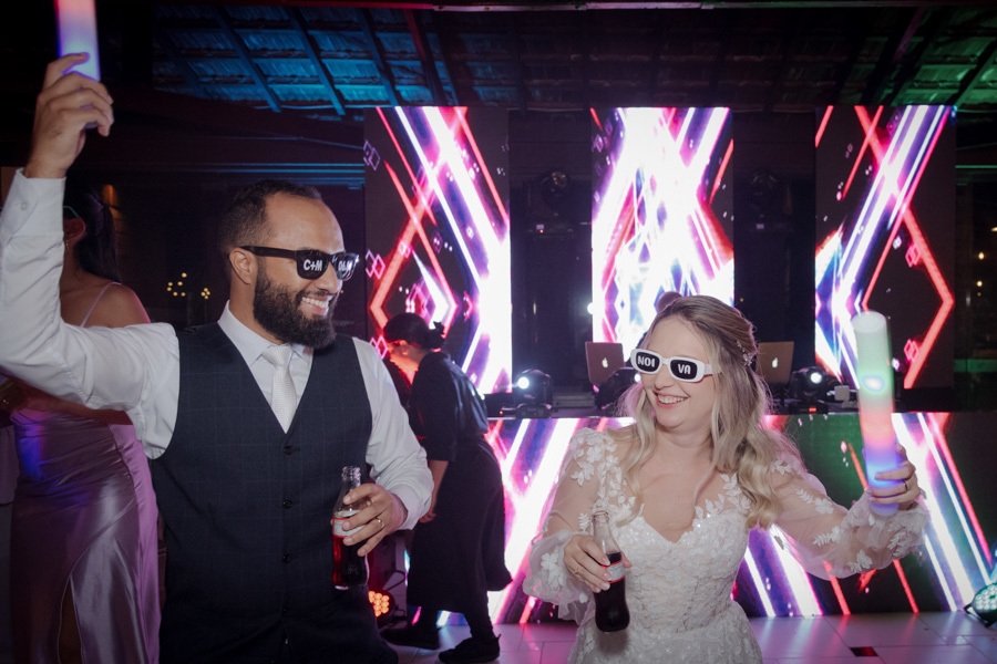 Noivos Claudia e Matheus curtem a festa com óculos personalizados e bastões de LED no Boulevard Jacareí. Fotografia de casamento animada com painel de LED ao fundo, capturando a descontração do casal, registrada pelo fotógrafo Gilson Dias.