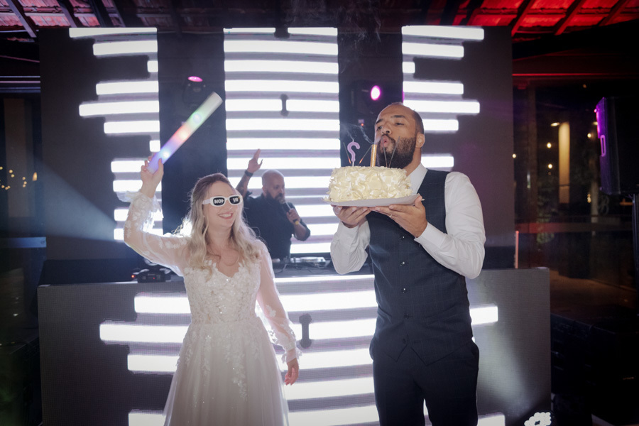 Noivo Matheus sopra a vela do bolo de casamento enquanto Claudia celebra com bastão de LED na pista de dança do Boulevard Jacareí. Fotografia de festa animada com painel de LED ao fundo e DJ, registrada com alegria pelo fotógrafo Gilson Dias.