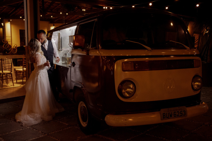 Noivos Claudia e Matheus se beijam ao lado de uma Kombi retrô de comida durante a festa no Boulevard Jacareí. Fotografia de casamento noturna capturando um momento romântico e charmoso na recepção, registrada pelo fotógrafo Gilson Dias.