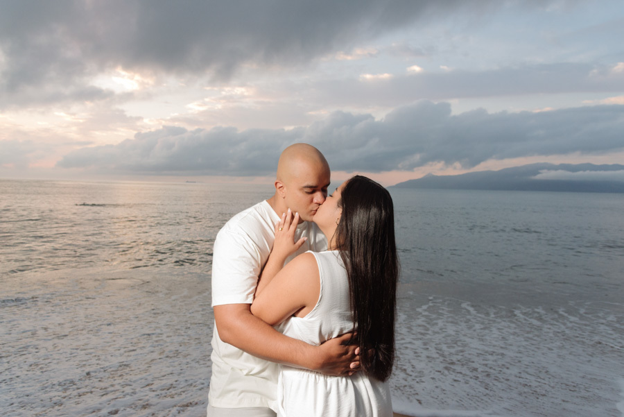 Fotografia de pré-wedding em Caraguatatuba: casal Stefanie e Alex se beijando na beira da praia ao amanhecer, vestindo roupas brancas com mar e montanhas ao fundo.