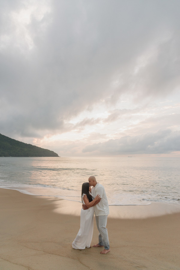 Fotógrafo em Caraguatatuba: Alex beija a testa de Stefanie em ensaio pré-casamento na areia. Plano aberto destacando a imensidão da praia ao amanhecer, com montanhas e céu nublado, transmitindo a serenidade do pré-wedding de Stefanie e Alex.