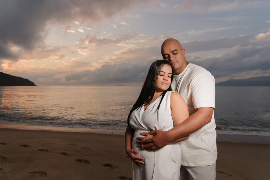 Fotografia de gestante e pré-wedding em Caraguatatuba: Alex abraça Stefanie por trás com as mãos na barriga, celebrando a espera pelo Heitor ao nascer do sol na praia. Um momento de conexão e amor em família capturado no Litoral Norte.