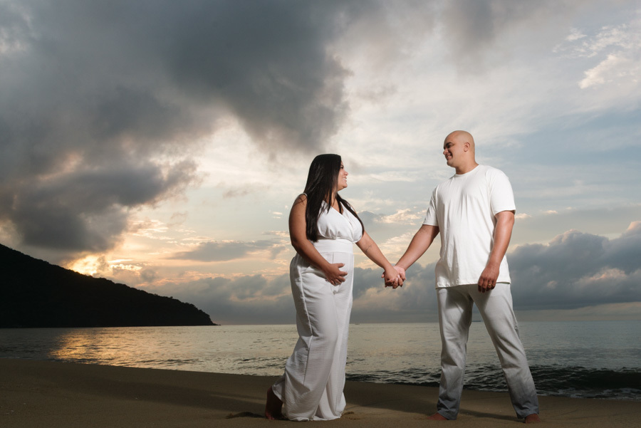 Ensaio pré-wedding em Caraguatatuba: Stefanie e Alex de mãos dadas na areia, olhando-se com cumplicidade ao nascer do sol. Fotografia de casal no Litoral Norte com céu dramático e luz suave, destacando a conexão e a espera pelo bebê Heitor.