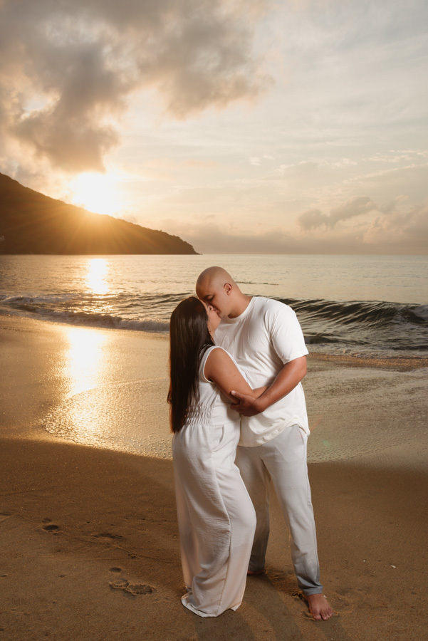 Fotografia pré-wedding em Caraguatatuba: Stefanie e Alex se beijam na praia com o sol radiante surgindo atrás da montanha. Ensaio de casal com luz dourada intensa e mar ao fundo, capturando o romantismo do nascer do sol no Litoral Norte.