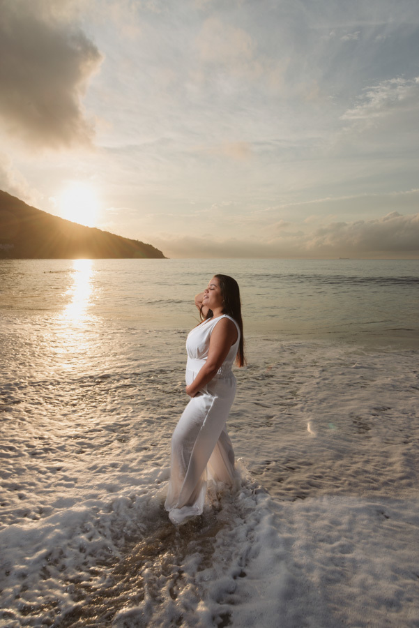 Ensaio gestante em Caraguatatuba: Stefanie posa na beira do mar com os pés na água ao nascer do sol. Fotografia profissional no Litoral Norte destacando a silhueta da grávida com luz dourada e mar ao fundo, aguardando a chegada do bebê Heitor.