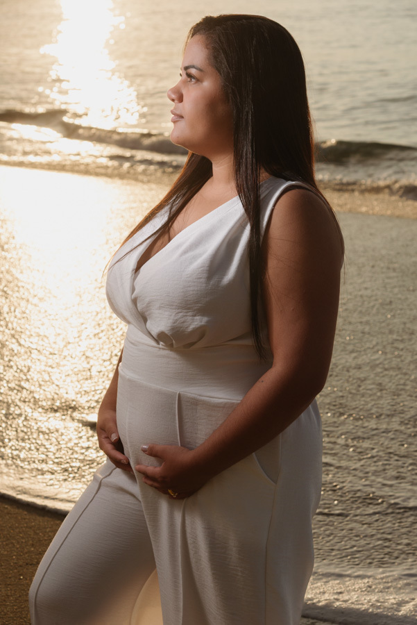 Foto de gestante em Caraguatatuba: Perfil de Stefanie com mãos na barriga e reflexo dourado do sol no mar ao fundo. Ensaio fotográfico profissional no Litoral Norte capturando a serenidade da espera pelo Heitor durante o amanhecer na praia.