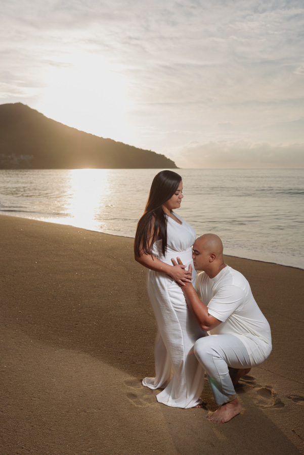 Fotografia de gestante em Caraguatatuba: Alex ajoelhado beija a barriga de Stefanie na areia da praia. Ensaio pré-wedding e família ao nascer do sol no Litoral Norte, registrando a doce espera pelo bebê Heitor com luz dourada e mar ao fundo.