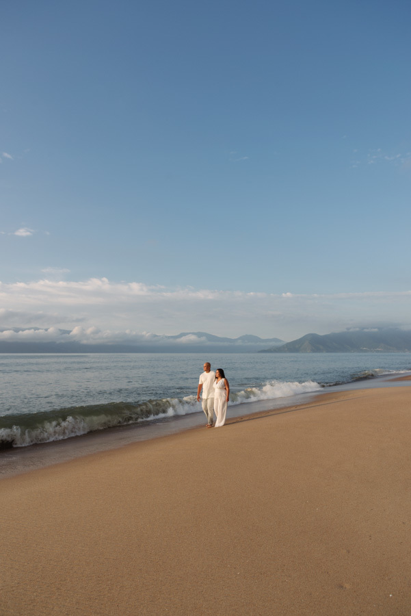 Ensaio pré-wedding em Caraguatatuba: Stefanie e Alex caminham na beira do mar sob um vasto céu azul. Plano aberto destacando a paisagem litorânea e a serenidade do casal no Litoral Norte, ideal para portfólio de fotografia de casamento e família.
