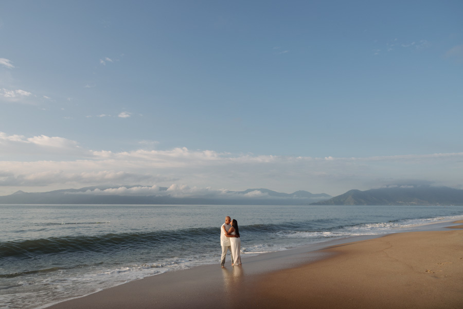 Ensaio pré-wedding em Caraguatatuba: Stefanie e Alex abraçados na beira do mar sob céu azul. Fotografia profissional com plano aberto destacando a paisagem do Litoral Norte e a conexão do casal, ideal para inspiração de casamento na praia.