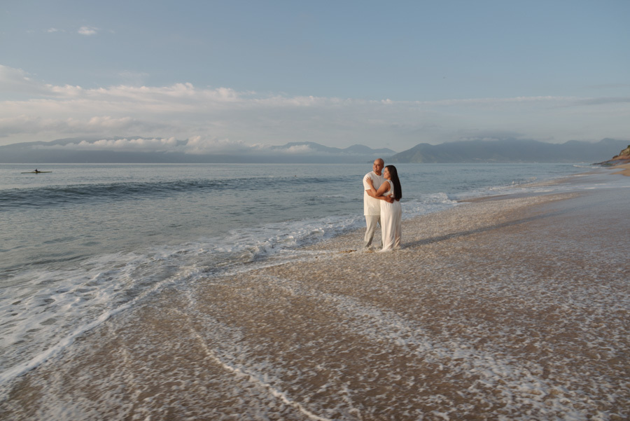 Ensaio pré-wedding em Caraguatatuba: Stefanie e Alex abraçados na beira do mar com espuma das ondas nos pés. Fotografia profissional no Litoral Norte com plano aberto, destacando a imensidão do oceano e montanhas ao fundo sob o céu claro da manhã.