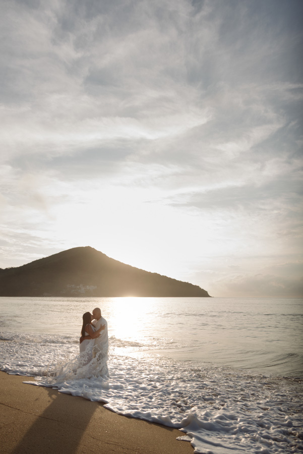 Ensaio pré-wedding em Caraguatatuba: Stefanie e Alex se beijam na beira do mar com a espuma das ondas nos pés. Fotografia com luz de contra-dia ao nascer do sol atrás da montanha, criando um cenário romântico e iluminado no Litoral Norte.