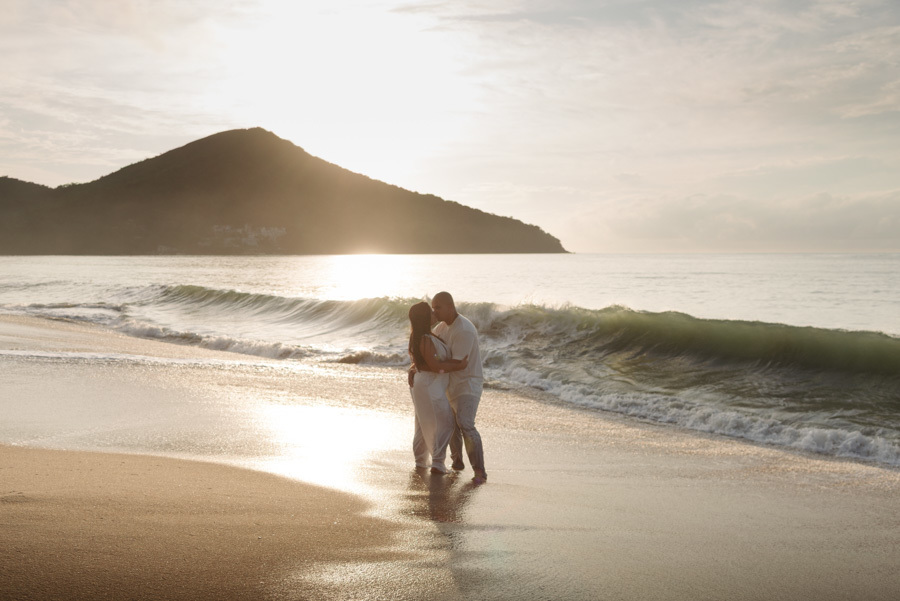 Ensaio pré-wedding em Caraguatatuba: Stefanie e Alex se beijam na beira do mar com uma onda quebrando ao fundo. Fotografia de casal no Litoral Norte com luz dourada do sol e montanhas, capturando um momento épico e romântico ao amanhecer.