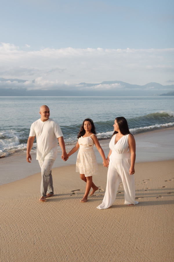 Ensaio de família em Caraguatatuba: Stefanie, Alex e a filha Elisa Vitória caminham de mãos dadas na areia da praia. Fotografia profissional no Litoral Norte capturando a união da família ao nascer do sol com mar e montanhas ao fundo.