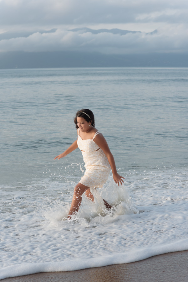 Fotografia de família em Caraguatatuba: Elisa Vitória brinca e corre na espuma das ondas durante ensaio no Litoral Norte. Captura espontânea e alegre ao nascer do sol, destacando a leveza e a celebração da vida na praia.