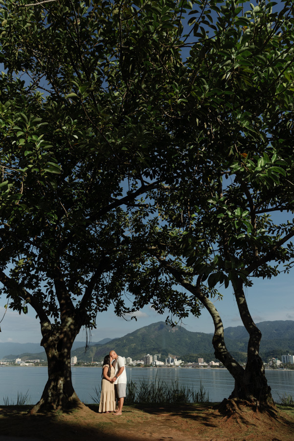 Fotografia pré-wedding em Caraguatatuba: Stefanie e Alex se beijam sob a sombra de árvores com a orla da cidade e montanhas ao fundo. Ensaio de casal no Litoral Norte destacando a silhueta, a natureza e a paisagem urbana de Caraguá.