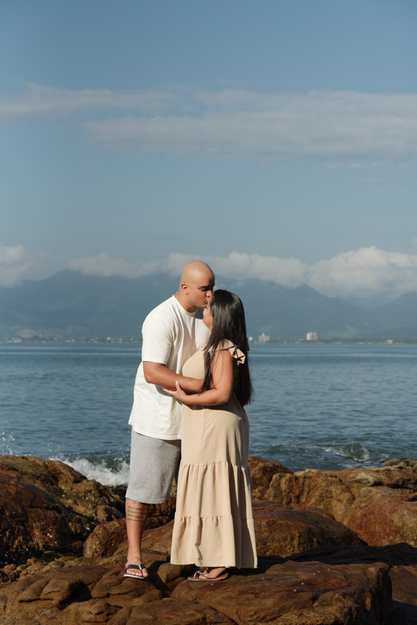 Fotografia pré-wedding em Caraguatatuba: Alex beija a testa de Stefanie sobre as rochas à beira-mar. Ensaio de casal no Litoral Norte com montanhas e céu azul ao fundo, destacando o romantismo e a cumplicidade do casal durante o dia.