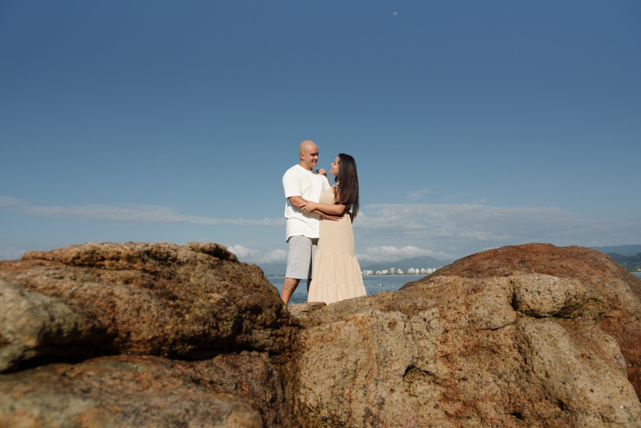 Ensaio pré-wedding em Caraguatatuba: Stefanie e Alex abraçados sobre rochas com mar e orla da cidade ao fundo. Fotografia profissional no Litoral Norte sob céu azul, destacando a conexão do casal e a paisagem litorânea em um dia ensolarado.