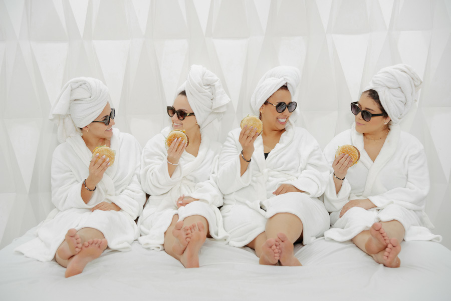 Noiva e madrinhas de roupão e óculos de sol comendo hambúrguer no making of em Taubaté. Foto divertida no Espaço Verde Vale, capturando momentos autênticos com fotografia de casamento espontânea por Gilson Dias no Vale do Paraíba.