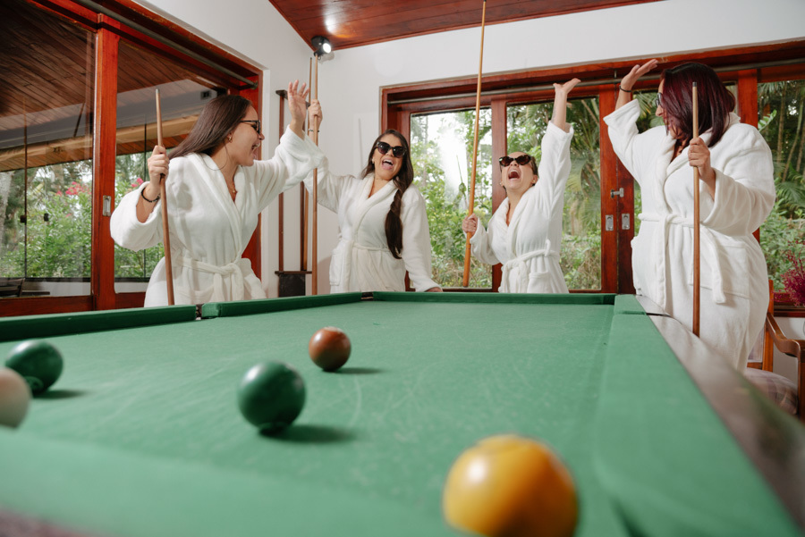 Noiva e madrinhas de roupão jogam sinuca e celebram no Espaço Verde Vale em Taubaté. Fotografia de making of descontraída e divertida no Vale do Paraíba, destacando momentos espontâneos antes do casamento capturados por Gilson Dias Fotografia.