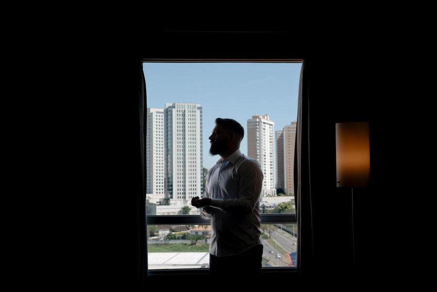 Silhueta do noivo se preparando em frente à janela com vista para São José dos Campos. Fotografia de making of masculina com luz e sombra, capturando a expectativa para o casamento no Vale do Paraíba pelo fotógrafo Gilson Dias.