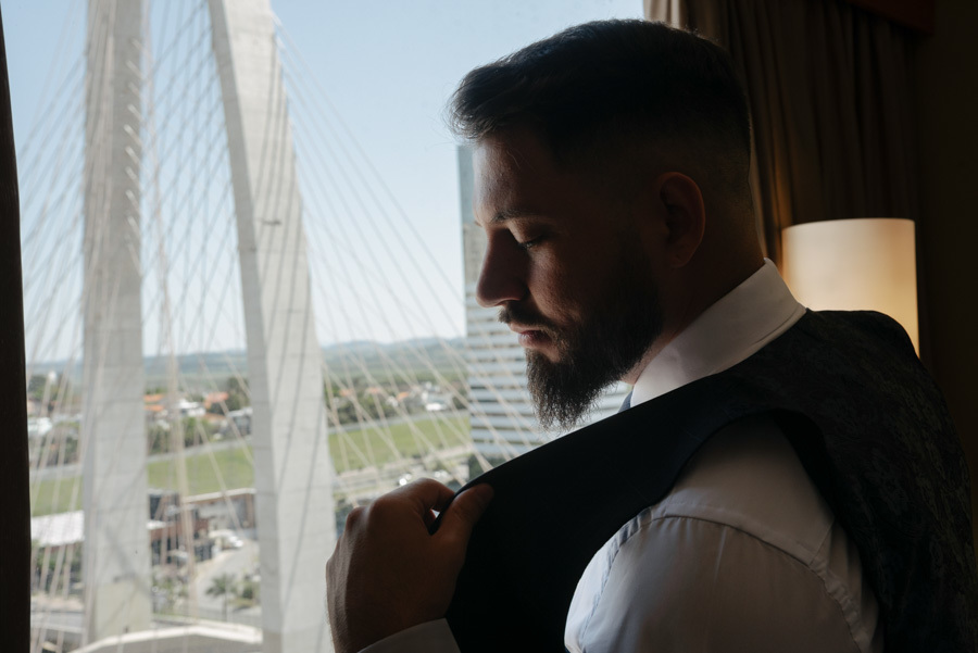 Close do noivo vestindo o colete durante o making of em São José dos Campos. Fotografia de casamento com foco nos detalhes e na expectativa do noivo, tendo ao fundo a Ponte Estaiada, registrada pelo fotógrafo Gilson Dias no Vale do Paraíba.