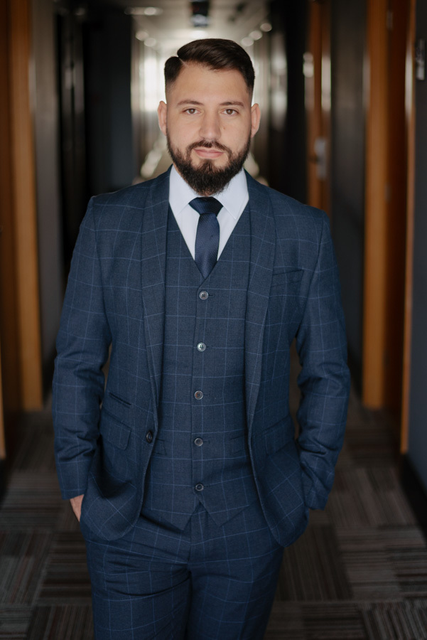 Retrato frontal do noivo Diego posando no corredor do hotel em São José dos Campos. Fotografia de casamento elegante destacando o terno xadrez azul, capturada pelo fotógrafo Gilson Dias como parte dos preparativos para o grande dia no Vale do Paraíba.
