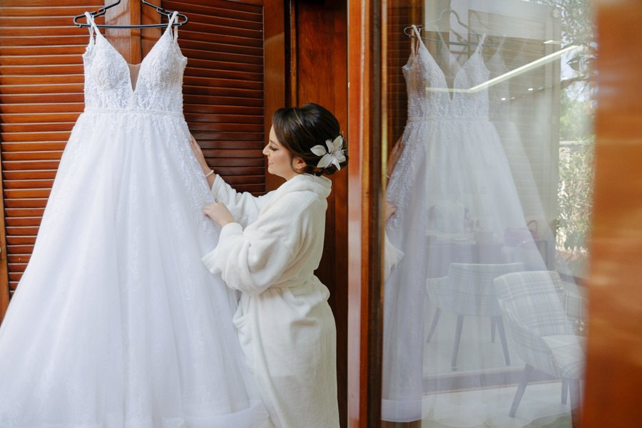 Noiva de roupão admira seu vestido de noiva branco pendurado durante o making of no Espaço Verde Vale, em Taubaté. Fotografia de casamento com foco na expectativa e detalhes da noiva, capturada por Gilson Dias no Vale do Paraíba.