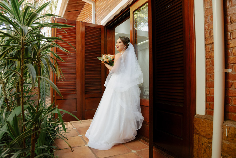 Noiva pronta sorri segurando o buquê na varanda do Espaço Verde Vale, em Taubaté. Fotografia de noiva iluminada e espontânea, destacando o vestido branco e o véu em meio à natureza, registrada pelo fotógrafo Gilson Dias no Vale do Paraíba.