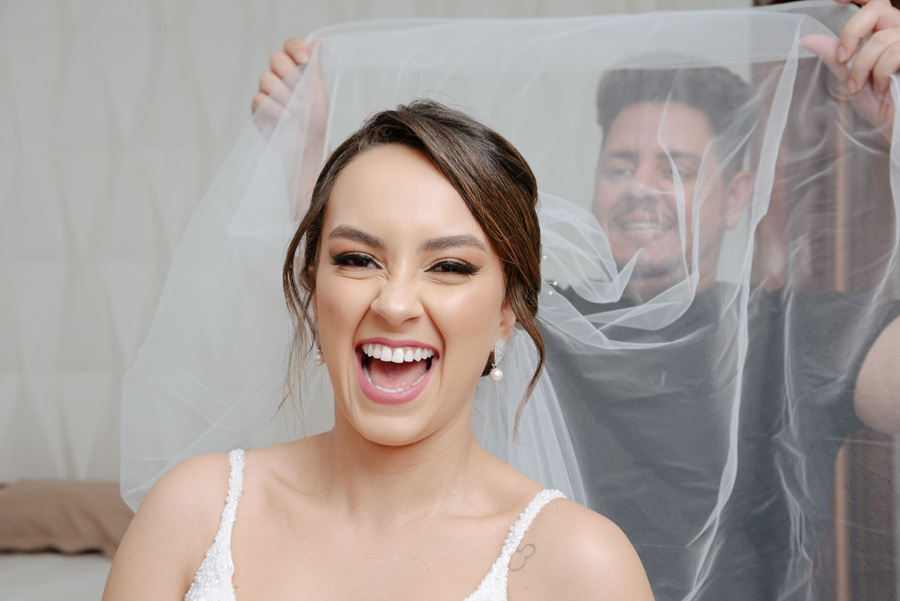 Noiva sorri alegremente enquanto o cabeleireiro ajusta seu véu durante o making of no Espaço Verde Vale, em Taubaté. Fotografia espontânea de casamento capturando a felicidade da noiva por Gilson Dias no Vale do Paraíba.
