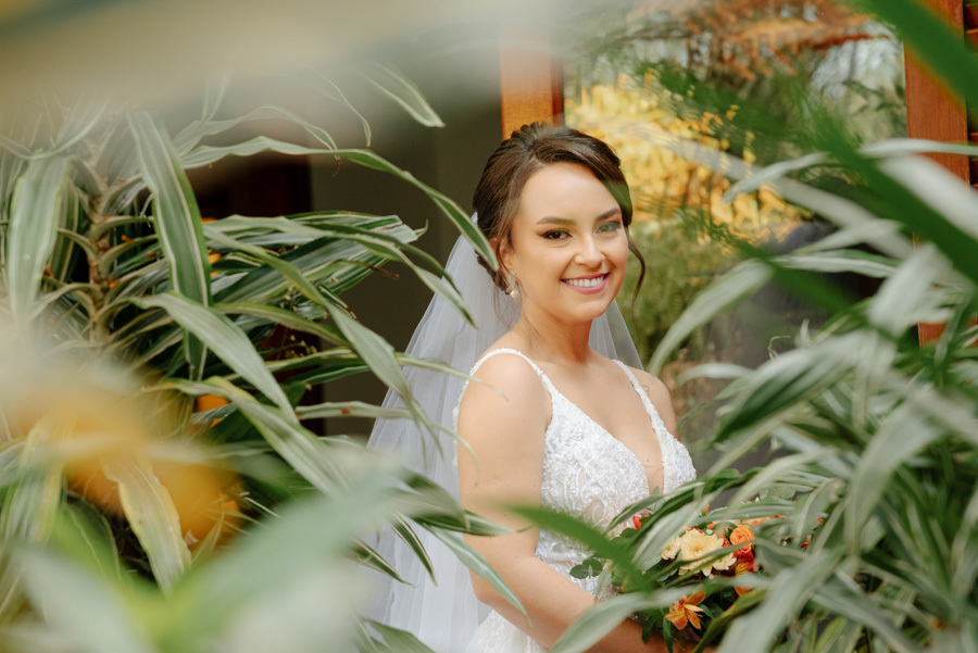 Noiva sorri em retrato emoldurado por folhagens no Espaço Verde Vale, em Taubaté. Fotografia de noiva com estilo natural e iluminado, destacando o buquê e o vestido branco em harmonia com a natureza, registrada por Gilson Dias no Vale do Paraíba.