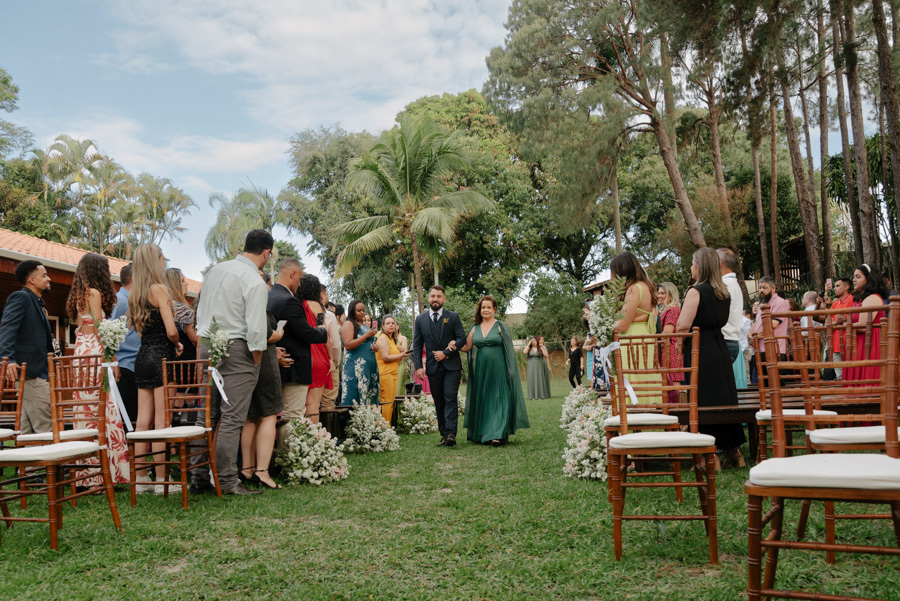 Noivo caminha até o altar acompanhado pela mãe em cerimônia ao ar livre no Espaço Verde Vale, Taubaté. Fotografia de casamento no Vale do Paraíba capturando o cortejo sob as árvores e o céu azul em um estilo natural por Gilson Dias.