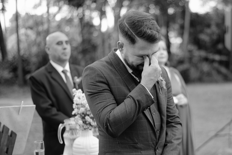 Noivo Diego chora de emoção no altar durante cerimônia ao ar livre no Espaço Verde Vale, em Taubaté. Fotografia de casamento em preto e branco capturando sentimentos reais e espontâneos no Vale do Paraíba, registrada por Gilson Dias Fotografia.