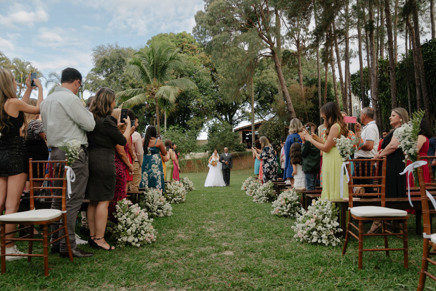Noiva Thayná inicia entrada na cerimônia ao ar livre no Espaço Verde Vale, Taubaté. Fotografia de casamento no Vale do Paraíba capturando o cortejo pelo gramado entre convidados e árvores altas, registrada pelo fotógrafo Gilson Dias.