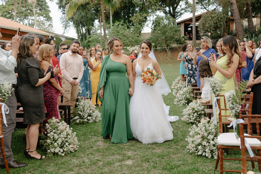 Noiva Thayná entra na cerimônia acompanhada pela mãe no Espaço Verde Vale, em Taubaté. Fotografia de casamento no Vale do Paraíba capturando a caminhada ao altar e sorrisos dos convidados sob luz natural, registrada por Gilson Dias Fotografia.