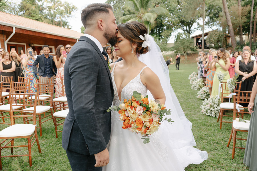 Noivo Diego beija a testa da noiva Thayná no altar durante cerimônia ao ar livre no Espaço Verde Vale, em Taubaté. Fotografia de casamento emocionante e espontânea no Vale do Paraíba, destacando o buquê laranja e o vestido de noiva, por Gilson Dias.