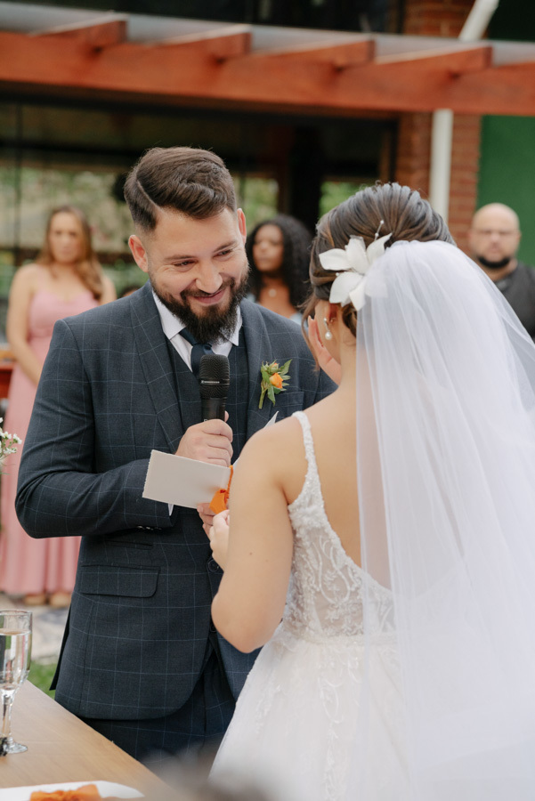 Noivo Diego sorri emocionado enquanto lê seus votos no microfone para a noiva Thayná. Fotografia de casamento em close capturando a cumplicidade do casal no altar do Espaço Verde Vale, em Taubaté, registrada pelo fotógrafo Gilson Dias.