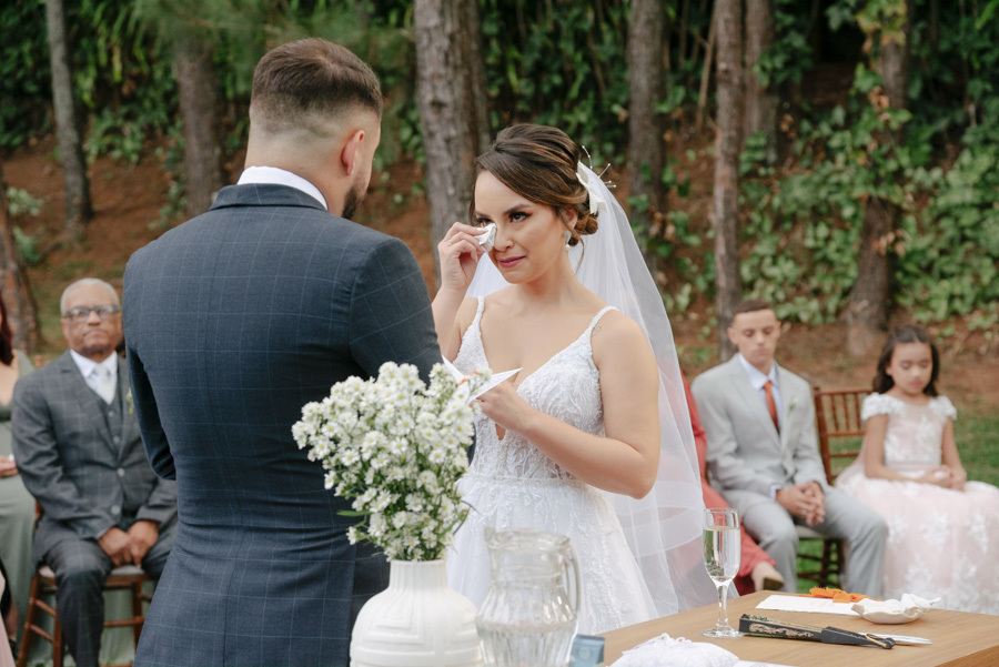 Noiva Thayná chora de emoção e enxuga as lágrimas durante a leitura dos votos no altar. Registro sensível de casamento ao ar livre no Espaço Verde Vale, em Taubaté, capturado pelo fotógrafo Gilson Dias no Vale do Paraíba.