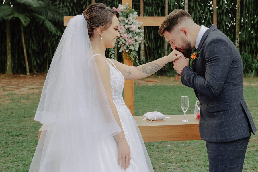 Noivo Diego beija a mão da noiva Thayná no altar durante a cerimônia no Espaço Verde Vale, em Taubaté. Momento romântico em frente à cruz de madeira decorada, registrado pelo fotógrafo Gilson Dias em estilo natural e iluminado no Vale do Paraíba.